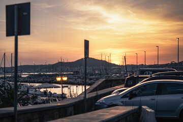 Beautiful evening cinematic photo, visible mountains, sunset, clouds and sky, red and yellow, sea, cars in shadow, parking lot, lanterns, many boats near the pier, water reflecting light.