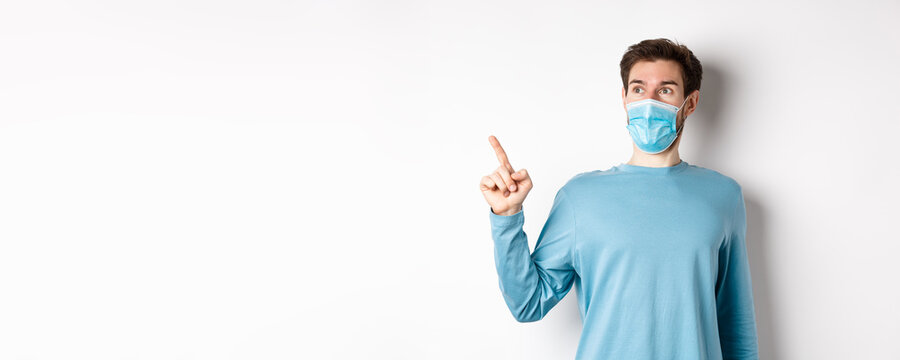 Coronavirus, Health And Quarantine Concept. Surprised Guy Looking In Awe And Pointing At Upper Left Corner, Standing In Medical Mask And Sweatshirt Over White Background