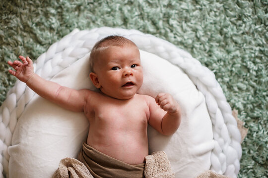 happy cheering naked newborn baby lying on a white round bed with arms up and fists looking into the camera as a winner shouting yay hooray