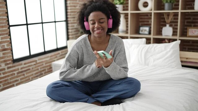 African american woman dancing and listening to music sitting on bed at bedroom