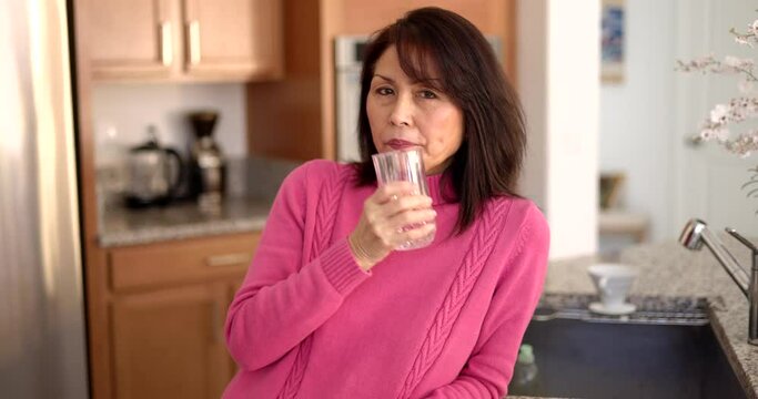 Attractive mature woman in pink sweater standing looking at camera smiling drinking glass of water in casual home kitchen at leisure home