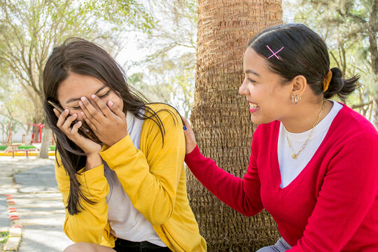 Mujer tap&aacute;ndose la cara mientras llama por tel&eacute;fono. Amigas conversando por llamada telef&oacute;nica en un parque.