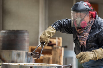 A female raku technician with red hair unloads ceramic ware with tongs, gloves and protective mask and helmet from a kiln