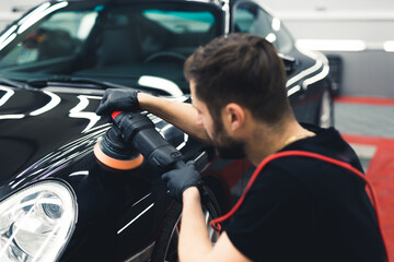 Rear view of white man crouching polishing the front of black car with professional buffer tool. Car detailing process. Horizontal indoor shot. High quality photo