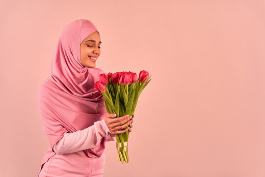  Spring And International Women's Day. A Young Beautiful Muslim Woman In A Soft Pink Hijab And Dress Is Holding A Bouquet Of Tulips And Looking At Them.