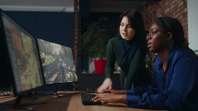 two female game designers discussing draft in office, black and caucasian ladies are looking at display
