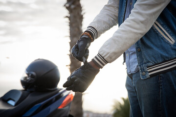 Hand of a biker putting on gloves with the scooter parked to the side with helmet on it.