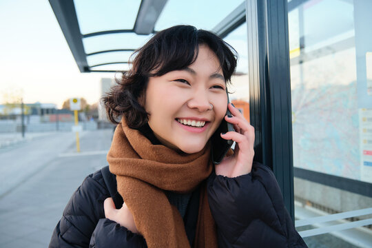 Close Up Of Cute Korean Woman, Making A Phone Call, Talking And Smiling On Telephone, Standing In Winter Jacket On Bus Stop, Waiting For Her Transport To Arrive