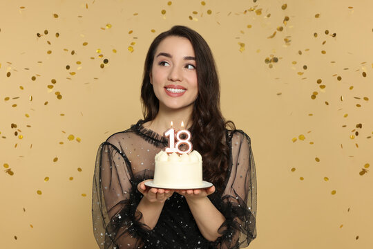 Coming of age party - 18th birthday. Woman holding delicious cake with number shaped candles and looking at falling confetti against beige background