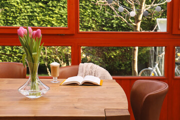 Glass of delicious cocoa, pink tulips and book on wooden table at terrace