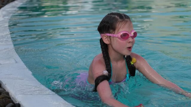 A cute little girl is swimming in an outdoor pool in swimming goggles and a purple swimsuit, diving and having fun. outdoors. The concept of family recreation.
