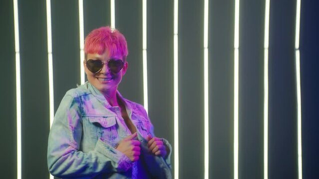 Active urban caucasian woman with short hot pink hair color in denim jacket dancing by neon lights background in disco club. Cool talented female dancer feeling free moves rhythmically hip-hop music