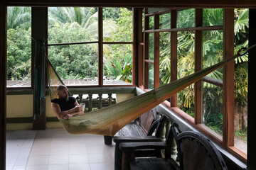 A tourist (middle aged man) is laying in a hammock with a cup or tea on the terrace of villa against jungle on rainy day. Krabi Province, Thailand.