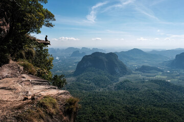 A tourist sits on the cliff edge overhanging the plain on sunny day. Dragon's Crest (Khao Ngon Nak) Viewpoint, Krabi Province, Thailand.