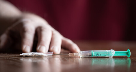 There is a syringe and drugs on a table. The hand of an anonymous person can be seen in the background.