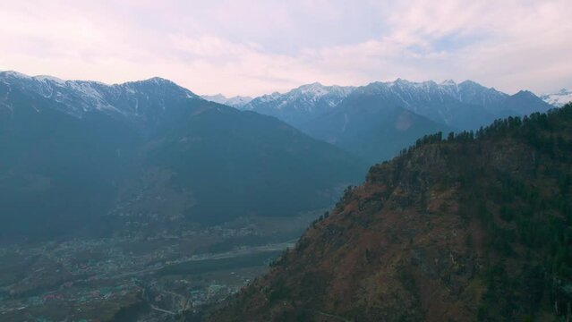aerial drone shot flying towards layers of mountains of himalaya range with tree covered hills and pink sky with rays showing manali town from hamta village a tourist spot