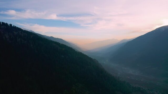 aerial drone shot flying towards layers of mountains of himalaya range with tree covered hills and pink sky with rays showing manali town from hamta village a tourist spot