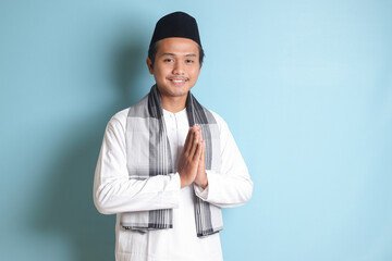Portrait of Asian muslim man in white koko shirt with skullcap showing apologize and welcome hand gesture. Isolated image on blue background - obrazy, fototapety, plakaty