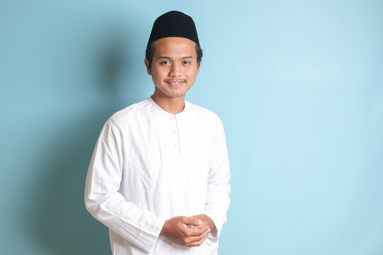 Portrait of religious Asian man in koko shirt or white muslim shirt and black cap, standing with crossed arms and looking at camera. Isolated image on gray background