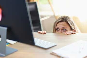 Scared female manager hidden behind office desk and looking at computer screen.
