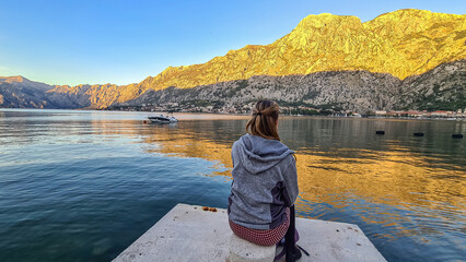 Woman sitting at pier with scenic view of Kotor bay at sunrise in summer, Adriatic Mediterranean Sea, Montenegro, Balkans, Europe. Fjord winding along coastal towns. First sunbeams on Lovcen mountains