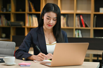 Positive focused Mature Business lady working with laptop at table in office, library with bookshelves on the background.