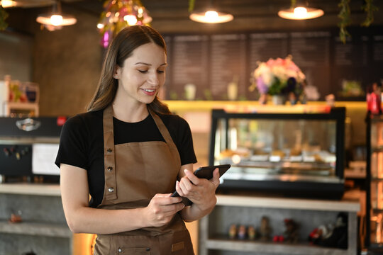 Beautiful caucasian woman business owner in apron standing in modern cafe interior and using digital tablet