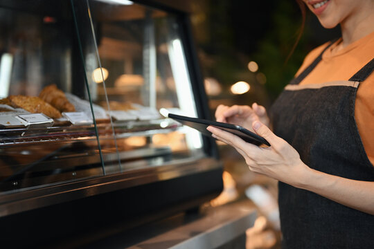 Smiling coffee shop owner using digital tablet to receiving online orders while standing near showcase with cakes and desserts