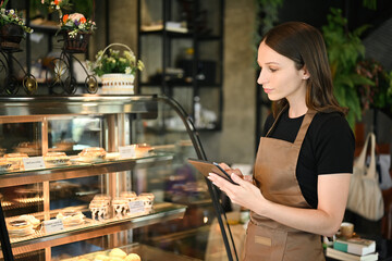 Caucasian female small business owner using digital tablet while standing near showcase with cakes and desserts