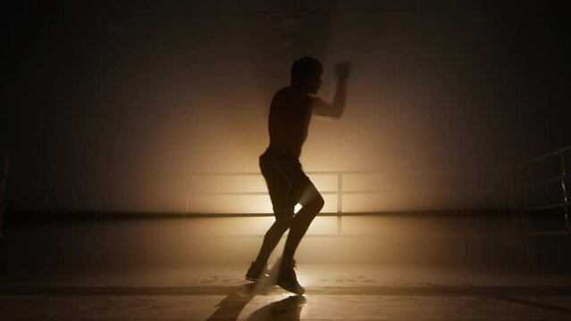 Silhouette of a kickboxer having an intensive workout in the gym. Unrecognized, strong man training in boxing club alone. High quality 4k footage in golden orange bronze foggy back light