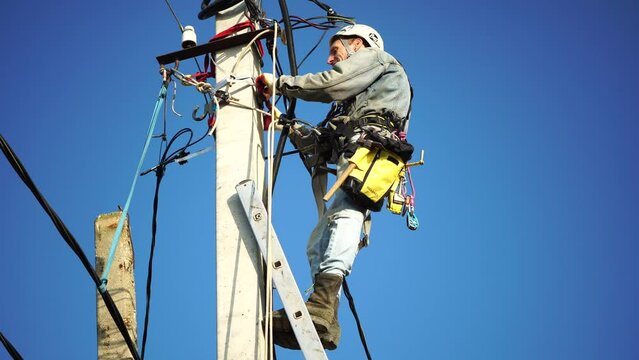 Skilled electrician in helmet fixes wires standing on ladder near high pole against blue sky on summer day backside view. Electrical service and mounting on the pole. Slow motion