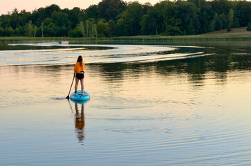 Fototapeta na wymiar Girl paddling on SUP board on beautiful lake during sunset or sunrise, standing up paddle boarding morning adventure in Germany lake district Mecklenburg 