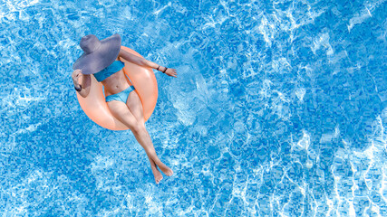 Beautiful woman in hat in swimming pool aerial top view from above, young girl in bikini relaxes and swims on inflatable ring donut and has fun in water on tropical vacation on holiday resort
