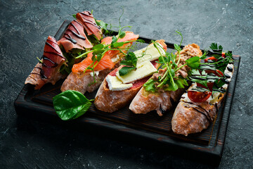 sandwich Set bruschetta with tuna, cheese, prosciutto and tomatoes on a dark wooden board. On a black stone background.