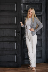 Vertical shoot of cheerful blonde Caucasian young woman in casual toothy smiles stands next to door home. Pretty model with folded hands in grateful looks at camera happily. Successful people.