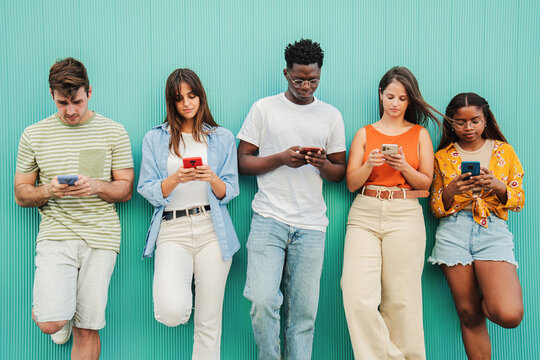 Group of multiethnic teenagers addicted to use the smartphone on blue background. Five multiracial young people or students, watching the screen of their cell phone and sharing media on a teal wall