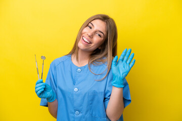 Dentist caucasian woman holding tools isolated on yellow background saluting with hand with happy expression