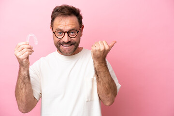 Middle age man holding invisible braces isolated on pink background pointing to the side to present...
