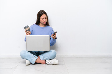 Young caucasian woman with laptop sitting on the floor isolated on white background holding coffee to take away and a mobile