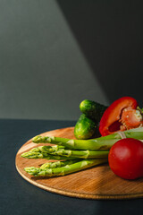 Vegetables lie on a wooden board: tomatoes, asparagus, cucumbers, red bell peppers. brown, dark gray background. place for text.