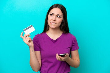 Young girl isolated on blue background buying with the mobile with a credit card while thinking