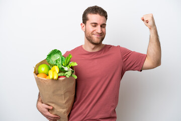 Young caucasian man holding a grocery shopping bag isolated on white background doing strong gesture