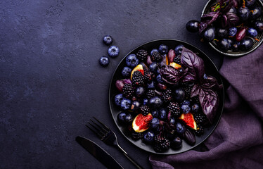 Summer blue and black berries fruit vegan salad: blueberries, blackberries, grapes, figs and purple basil with honey dressing on dark background, top view