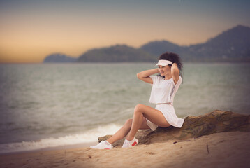 Holiday portrait of a pretty young brunette model with long sexy muscular legs and curly hair dressed in a white skirt, shirt, sneakers and white sun visor  sitting on the timber stump at the beach.