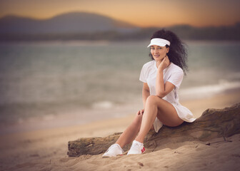 Fototapeta premium Holiday portrait of a pretty young brunette model with long sexy muscular legs and curly hair dressed in a white skirt, shirt, sneakers and white sun visor sitting on the timber stump at the beach.