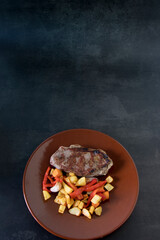 Entrecot con guarnición de patata, boniato y pimiento rojo, en un plato de cerámica marrón sobre mesa de mármol negro.