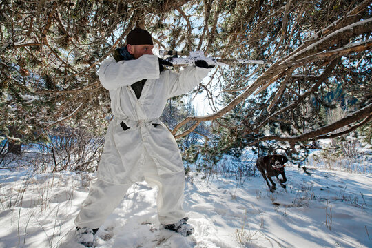 A hunter wearing  a cold-weather camouflage outfit aims his rifel in preparation to shoot while his hunting dog prepares to retrieve.