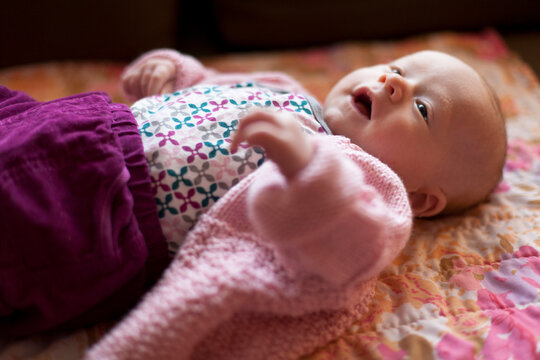 A newborn baby sits on her back on a couch smiling and looking up in a home.