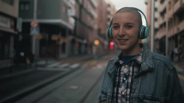 Shaved head girl listening to music with headphones while waiting for public transportation in the city street