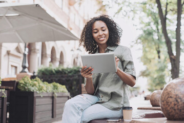 Smiling young woman using digital tablet in a city. Beautiful student girl having coffee break. Modern lifestyle, connection, online business education, freelance work concept
